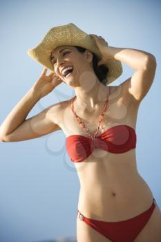 Caucasian mid-adult female in swimsuit holding hat on head laughing.