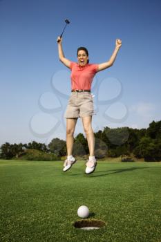Caucasion mid-adult woman holding golf club jumping in air cheering with golf ball and hole in foreground.