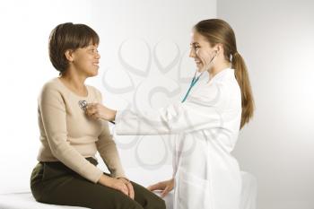 Caucasian mid-adult female doctor examining African American middle-aged female patient with stethoscope.