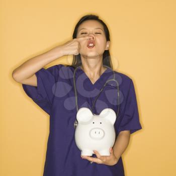 Asian Chinese mid-adult female doctor holding piggy bank against yellow background and touching nose.