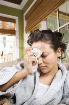 Ill woman in a bathrobe holds tissue to her forehead. Vertical shot.