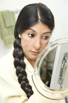 A young woman wearing a bathrobe is sitting in front of a mirror and applying makeup. Her long dark hair is braided and hanging over her shoulder. Vertical shot.