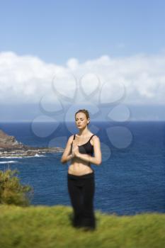 Attractive young woman stands with eyes closed in meditation in a green field overlooking the coast. Vertical shot.