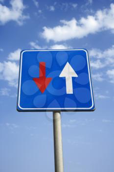 Low angle view of road sign with arrows in Tuscany, Italy. Blue sky and clouds are in the background. Vertical shot.
