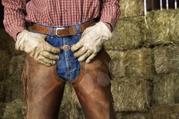 Close up of man with hands on his waist, wearing chaps and standing in front of hay bales. Horizontal shot.