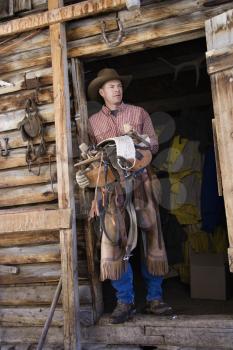 Man wearing a cowboy hat, leaning in the doorway of log cabin and holding a saddle. Vertical shot.