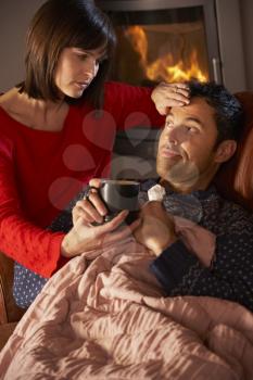 Wife Nursing Sick Husband With Cold Resting On Sofa By Cosy Log Fire