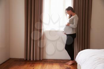 Pregnant Woman Standing By Window In Bedroom Holding Belly