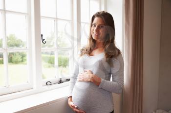 Portrait Of Pregnant Woman Standing By Window Holding Belly