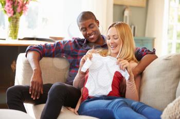 Pregnant Couple On Sofa At Home Looking At Baby Clothes