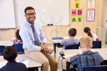 Smiling elementary school teacher sitting on a pupils desk