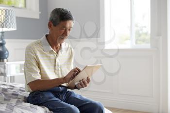 Senior Man Sitting On Bed Using Digital Tablet