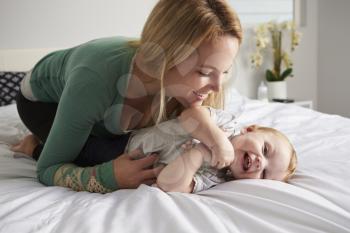 Caucasian mother kneeling on bed, tickling her baby daughter