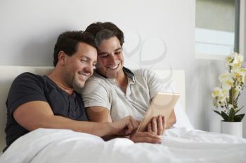 Male gay couple relax in bed together using digital tablet