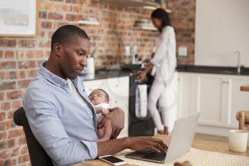 Father On Laptop Holds Newborn Son As Mother Makes Meal