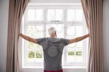 Senior Man Opening Bedroom Curtains And Looking Out Of Window