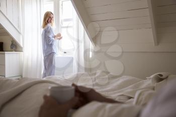 Woman Opens Curtains And Looks Out Of Window As Man Lies In Bed