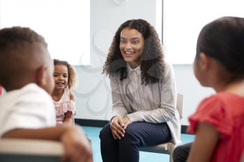 Infant school children sitting on chairs in a circle in a classroom talking to their smiling female teacher, close up, selective focus