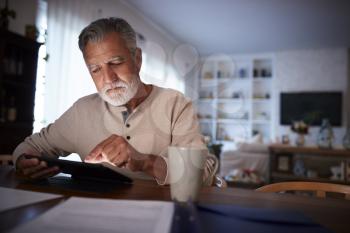 Senior Hispanic man sitting at his dining table reading an e book at home in the evening, close up