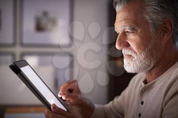 Senior Hispanic man sitting at a table using a tablet computer  in the evening, close up, side view