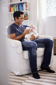 Smiling Hispanic father holding his four month old son feeds him a bottle, full length, vertical