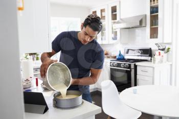 Millennial Hispanic man pouring cake mixture into cake form, following a recipe on a tablet computer