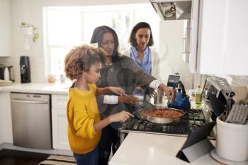 Pre-teen black girl standing at the hob in the kitchen preparing food with her grandmother and mother, selective focus