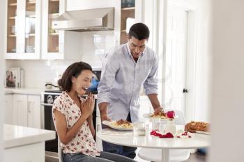 Young mixed race adult woman sitting at the table in the kitchen, her partner surprising her by serving a romantic meal, selective focus