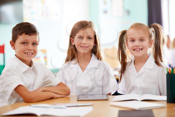 Portrait Of Three Elementary School Pupils Wearing Uniform Using Digital Tablet At Desk