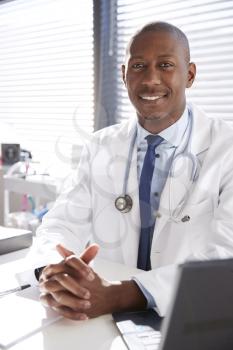 Portrait Of Smiling Male Doctor Wearing White Coat With Stethoscope Sitting Behind Desk In Office