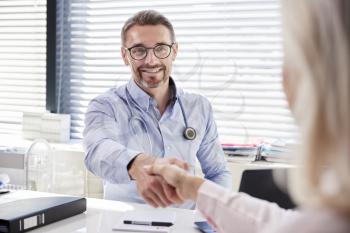 Female Patient Shaking Hands With Doctor Sitting At Desk In Office