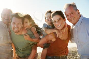 Three generation white family on a beach smiling to camera, parents piggybacking kids, close up