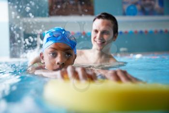 Male Swimming Coach Giving Boy Holding Float One To One Lesson In Pool