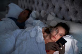 Woman Lying In Bed Checking Mobile Phone Whilst Man Sleeps Next To Her