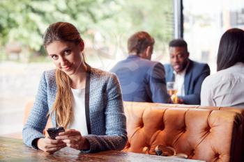 Portrait Of Businesswoman Sitting In Bar Checking Mobile Phones Whilst Colleagues Meet For Drink