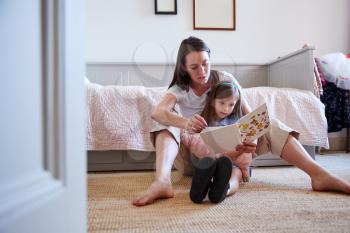Mother Sitting On Bedroom Floor Reading Book With Daughter At Home Together