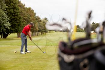 Mature Male Golfer Preparing To Hit Tee Shot Along Fairway With Bag Of Clubs In Foreground