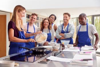 Female Teacher Making Pancake On Cooker In Cookery Class As Adult Students Look On