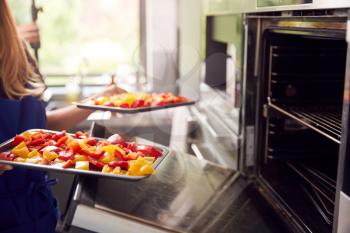 Close Up Of Woman Wearing Apron In Kitchen Putting Trays Of Peppers For Roasting In Oven
