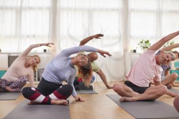 Group With Teacher Sitting On Exercise Mats Stretching In Yoga Class Inside Community Center