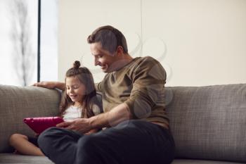 Father And Daughter Sitting On Sofa At Home Playing Together On Digital Tablet In Pink Case At Home