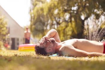 Mature Man Lying On Grass And Sunbathing At Home As Children Play In Pool