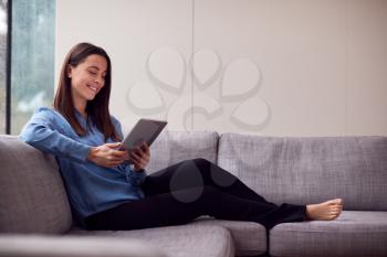 Smiling Young Woman At Home Sitting On Sofa Looking At Digital Tablet