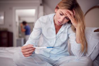 Unhappy Woman Wearing Pyjamas In Bedroom Holding Pregnancy Test