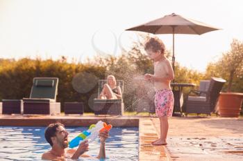 Father Squirting Son With Water Pistol Playing In Swimming Pool On Summer Vacation