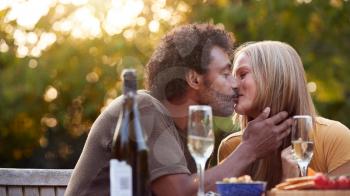 Mature Couple Kissing As They Celebrate With Champagne Sitting At Table In Garden With Snacks