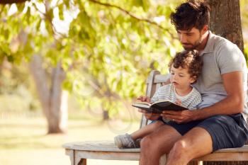 Father Sitting On Park Bench Under Tree With Son Reading Book Together