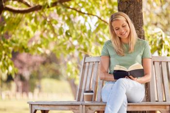 Woman Sitting On Park Bench Under Tree Reading Book And Drinking Takeaway Coffee