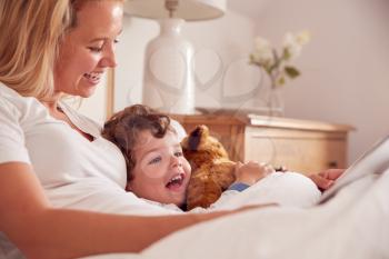 Mother In Bedroom Looking At Picture Book With Young Son Wearing Pyjamas Cuddling Soft Toy