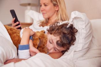 Young Son Cuddles Teddy Bear In Parents Bed Whilst Mother Looks At Mobile Phone